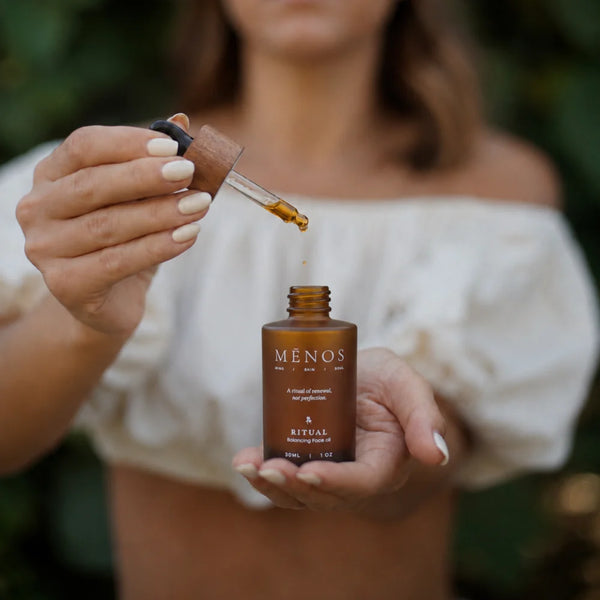 Person holding a MĒNOS RITUAL face oil bottle beneath a dropper of golden oil